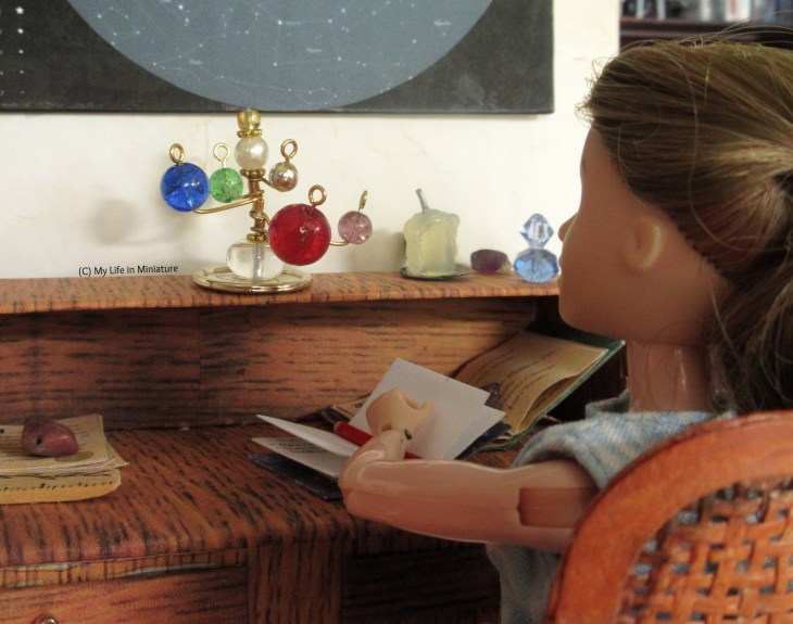 Sarah sits at her desk in her room, one hand holding a notebook open. The shot looks over her shoulder as she looks at the orrery, or solar system model, on the desk's shelf. The orrery has five planets of various colours on wires, circling a central pearlescent bead.