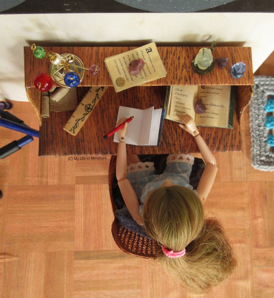 Overhead shot of Sarah's desk, as she is sitting at it to write. At desk level is also a large astronomy book, held open with a coloured gemstone, a ruler, and various scrolls and papers. On the shelf is the orrery, a stack of papers weighted down with another gemstone, a candle, another gemstone, and an inkjar.