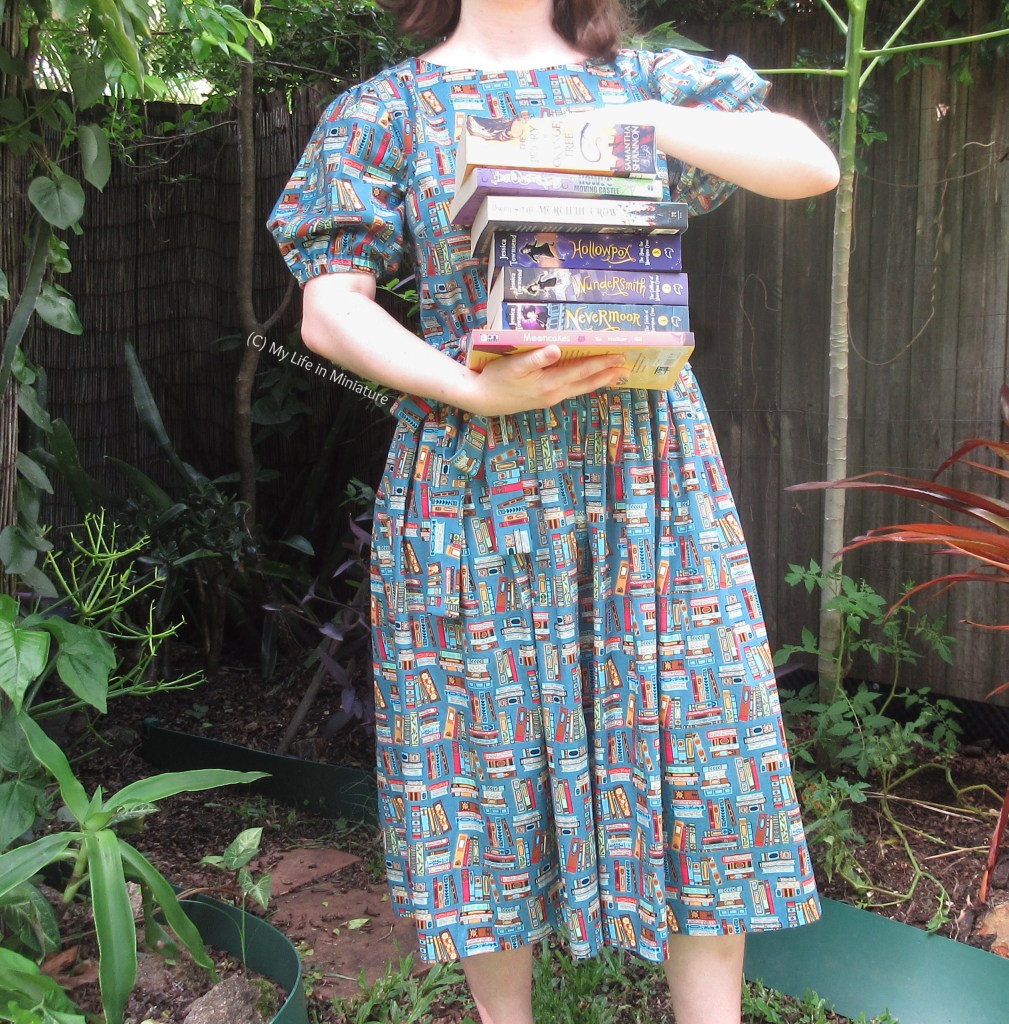 The author is photographed frmo shoulders to ankles, still wearing the book dress and under the leafy archway. She holds a big stack of novels in front of her torso, one hand supporting from below and one stabilising from above. 