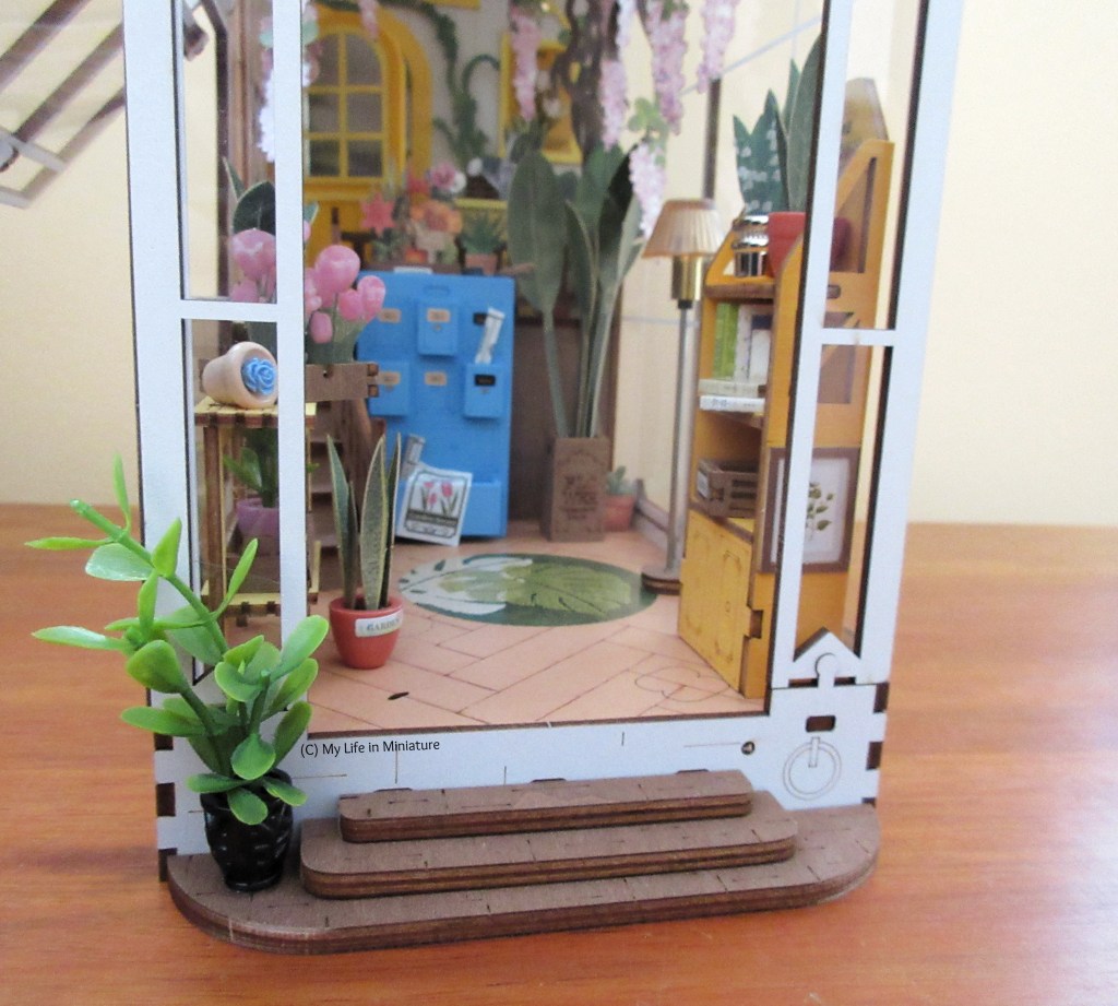 Close-up of the greenhouse book nook interior. The rocking chair and some smaller items have been removed, so the interior is easier to see. There are small tables, shelving units, chests of drawers all packed with pot plants, plant books, plant posters, etc. There are some pot plants on the floor, one on the step outside the greenhouse entrance, and a free-standing floor lamp.