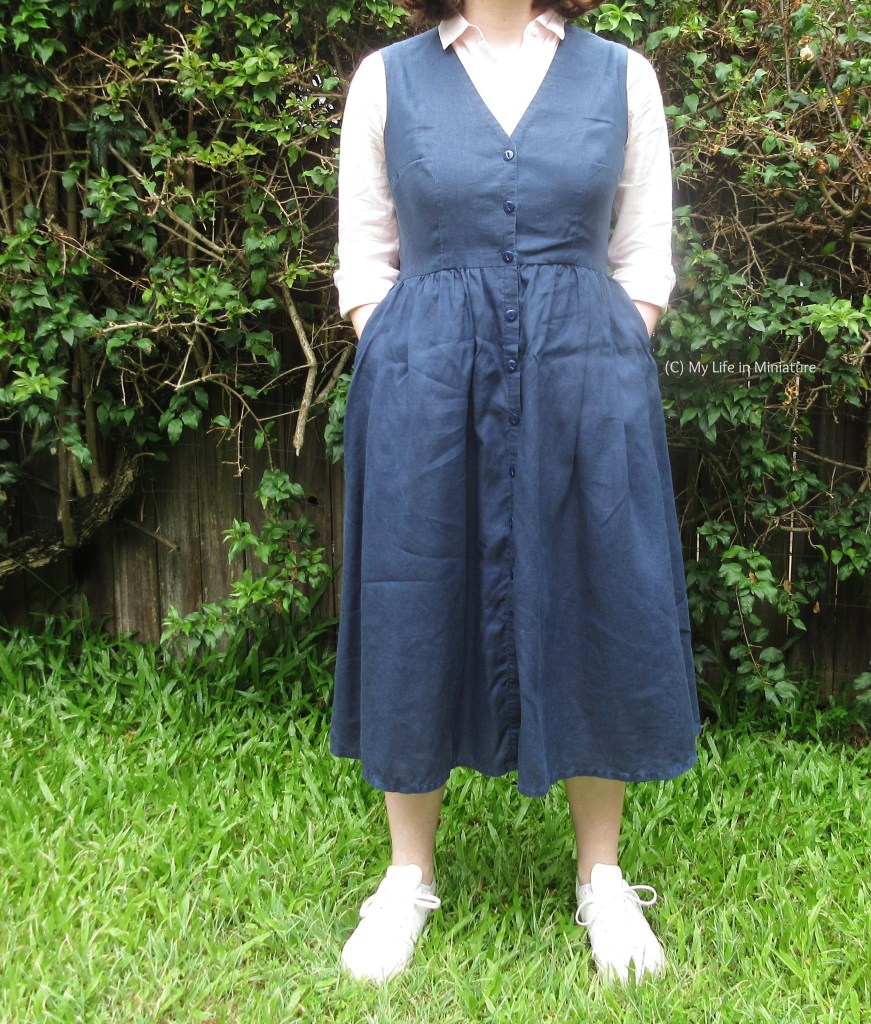 The author stands in front of a hedge, photographed from the neck down. She wears the navy pinafore and pink button-down from before, this time with her hands in the side-seam pockets of the skirt. She also has white shoes on. 