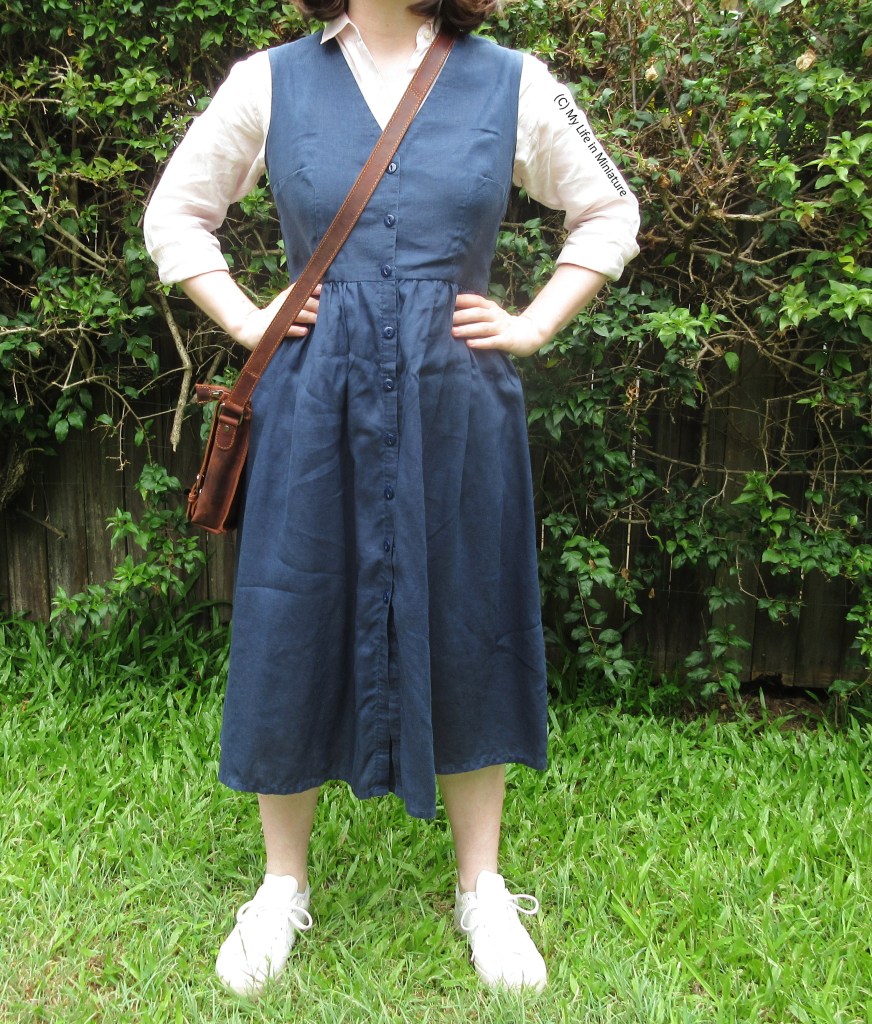 The author stands in front of a hedge, facing the camera and photographed from the shoulders down. She wears a navy V-necked pinafore dress over a pale pink button-down shirt, with a brown leather cross-body bag at her hip. Her hands are on her hips. 