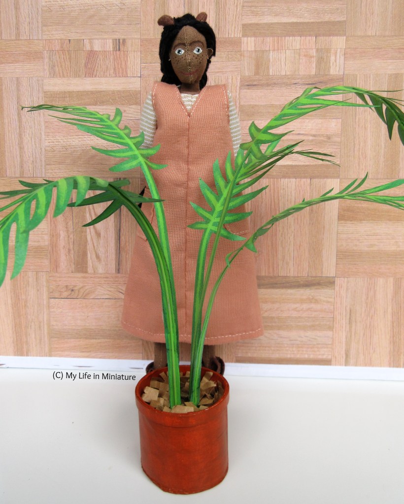 Petra stands behind a potted palm, smiling at the camera with her hands behind her back. She wears a peach pinafore dress with a cream-and-white striped top. The palm reaches her shoulders, and is in a terracotta pot. 