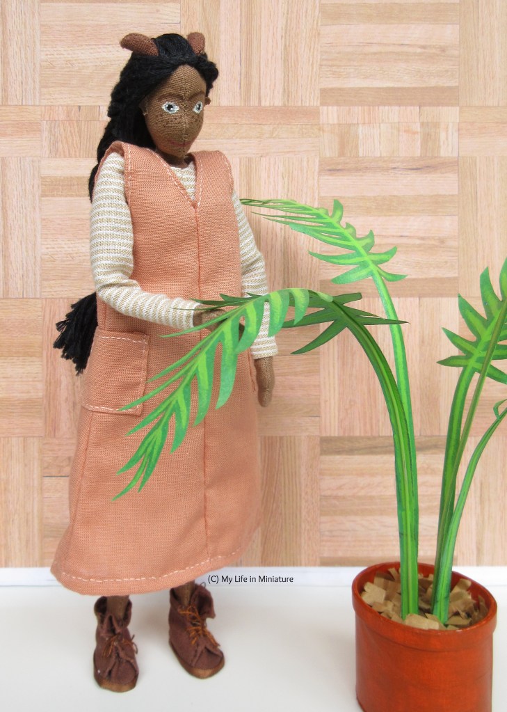 Petra stands in front of a parquet background, next to her potted palm. She supports one of the leaves with her hand from beneath, and she looks down at this leaf. With her pinafore and shirt, she has brown boots. 