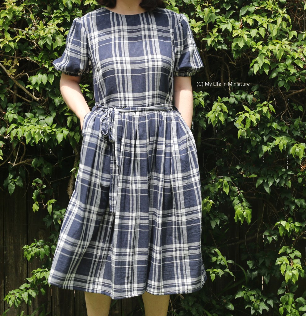 The author stands outside in front of a hedge. She faces the camera, wearing a navy and white checked dress. She has her hands in her pockets. She is photographed from neck to ankles. 