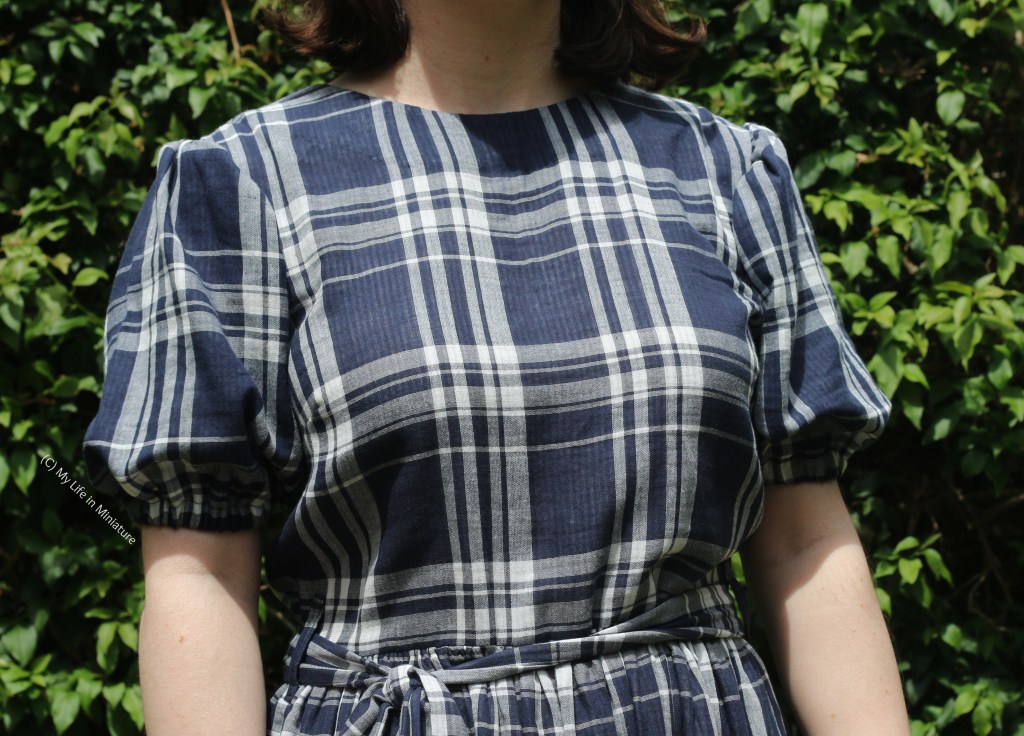 Close-up of the bodice of the checked dress. The checks are white on the navy background. There are puffed sleeves, a matching tie belt, and a circular neckline. 