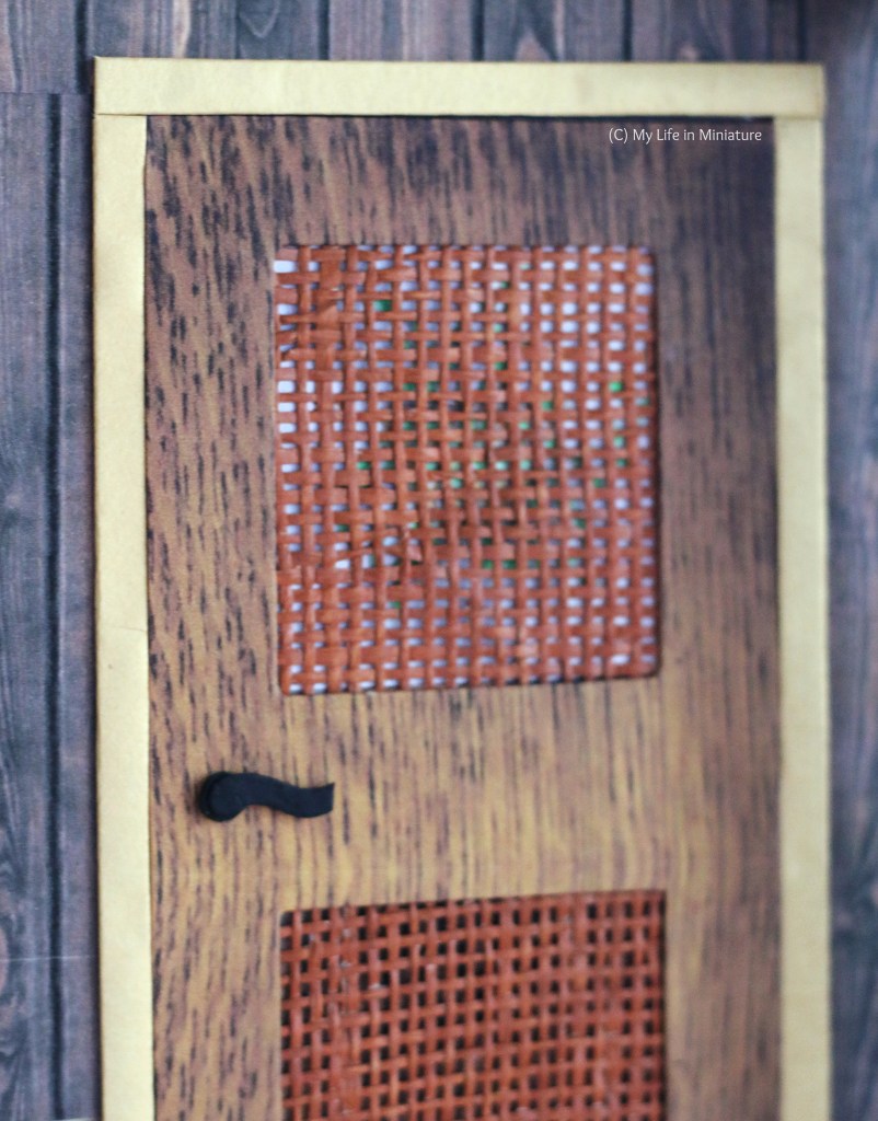 Close-up of the top half of the door to Tea at Yaz's. The door is wood, with a light gold frame. Panels in the door are latticed, and something green is just visible behind the top panel's lattice. There is also a black door handle. 