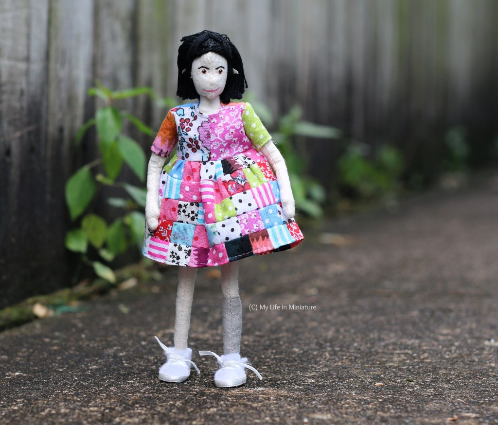 Tiffany stands outside, with a grey fence and some weeds in the background. She faces the camera, hands by her sides, smiling down at her dress. Said dress is completely mis-matched; a patchwork of many different fabrics of all colours and prints. It comes to just above her knees, and she has white shoes on. 
