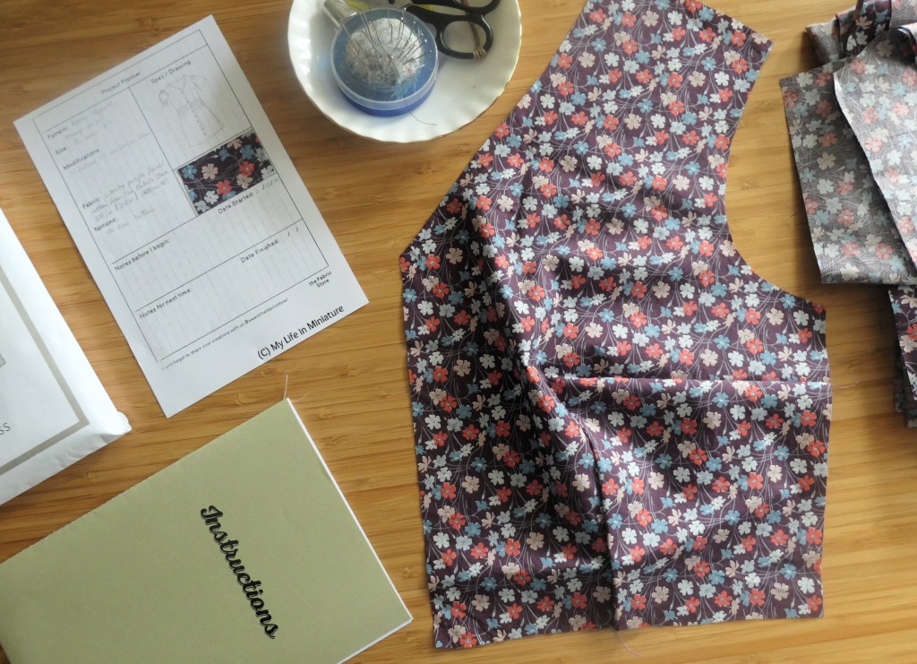 A flatlay of an assortment of sewing-related objects, on a wood background. Such objects include the front bodice of a dress, a dish with pins, scissors, and suchlike in, an instruction booklet, a project planning sheet, and the corner of a pattern pouch. The fabric is purple with red, pink, blue, and white flowers all over. 