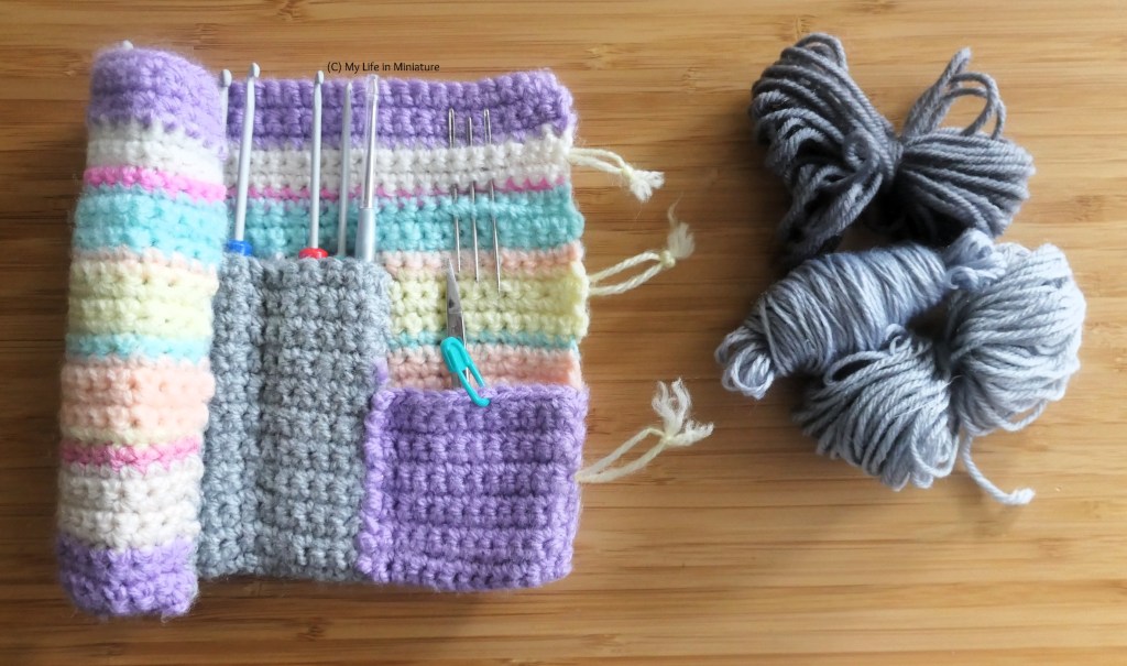 The author's crochet hook roll sits partially unrolled beside three small skeins of grey acrylic yarn, all on a wood surface. 