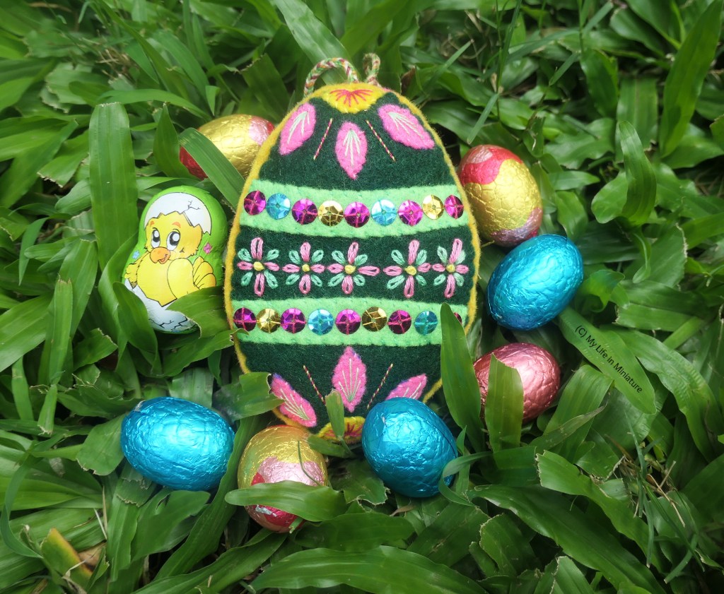The egg sits in grass, surrounded by small chocolate eggs in pink, blue, and gold. 