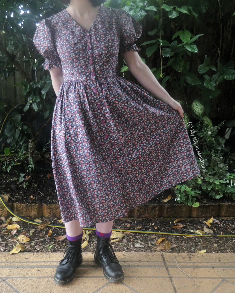 The author holds out the skirt of the purple dress, still in front of the greenery wall. 