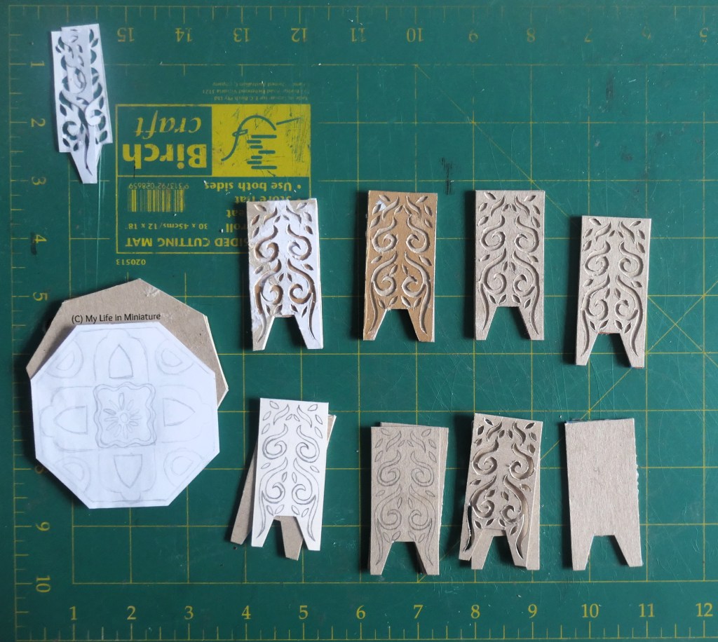 Overhead shot of the parts of a miniature table, laid out on a green cutting mat. There are eight rectangles like the one Petra had, two of which haven't had the designs cut out yet. To their left, there is an octagonal piece of cardboard under an octagonal piece of paper. The paper has a rough, symmetrical design on it. 