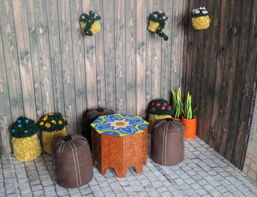 Wide shot of a corner of Tea at Yaz's courtyard dining area. In this corner are multiple potted plants, a couple of hanging baskets, and a table with thtee brown poufs around it. The table is octagonal, has carved wooden legs, and a brightly patterned surface. It's roughly the same height as the poufs. 