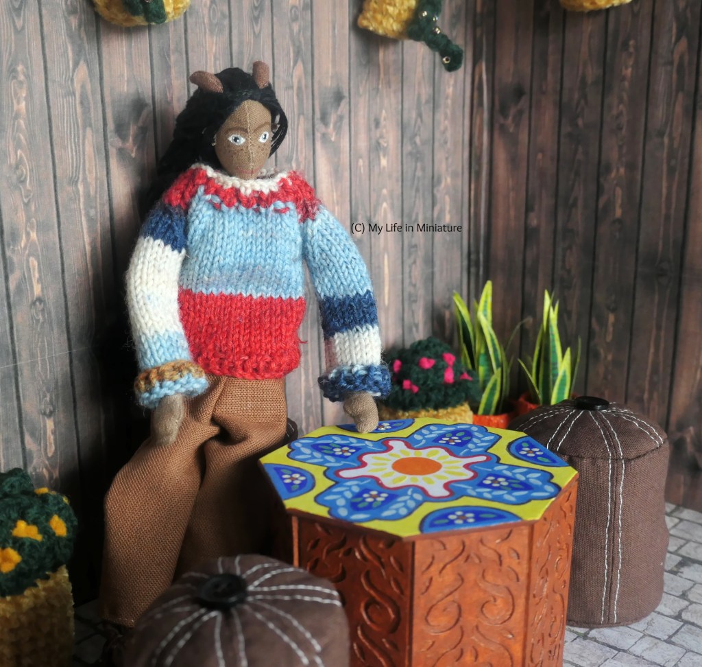 Petra sits at the table in Tea at Yaz's courtyard. She has one hand on her knee and one gently running over the surface of the table. She's looking down at the table, wearing a multi-coloured knit jumper and brown pants. 
