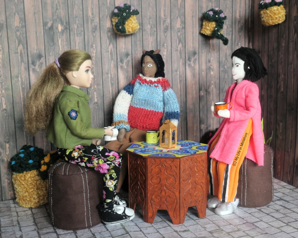 Petra, Tiffany, and Sarah sit around the table in Tea at Yaz's courtyard. They all have mugs with hot drinks in their hands, and they look to Sarah as she says something. There is a small gold lantern in the centre of the table, unlit.