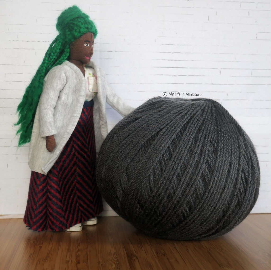 Hazel stands beside one of the balls of grey yarn, with one hand resting on it. The ball reaches to about her waist in height. 