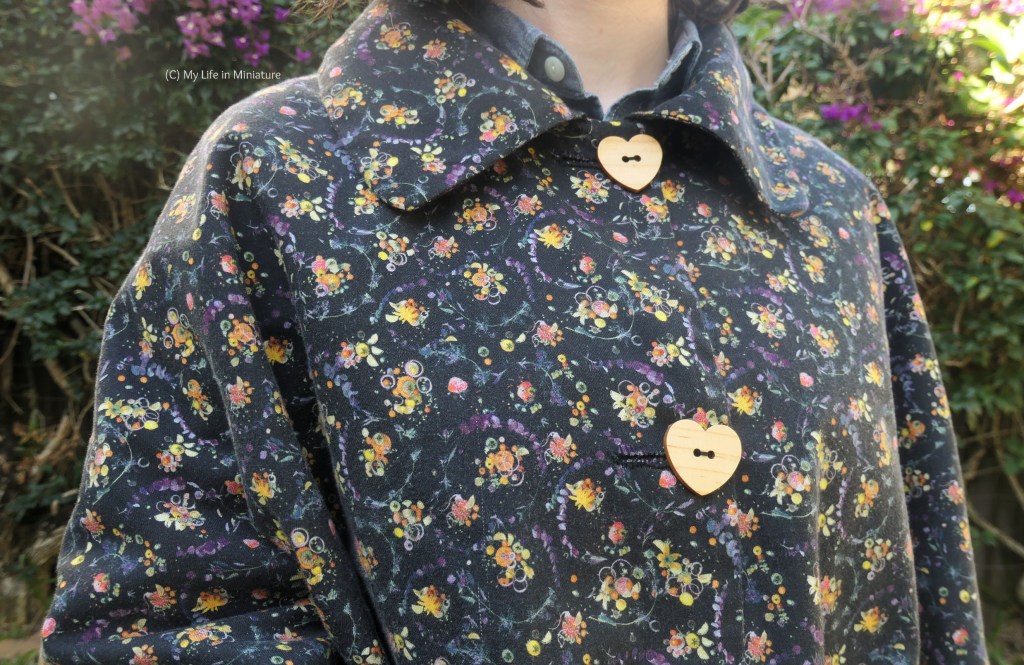 Close-up of the front of the cape. The large collar has curved front corners, and the two wooden buttons are in the shape of hearts. The cape fabric has pink, yellow, and orange berries in small clusters, interspersed with purple flowers, against a black background.