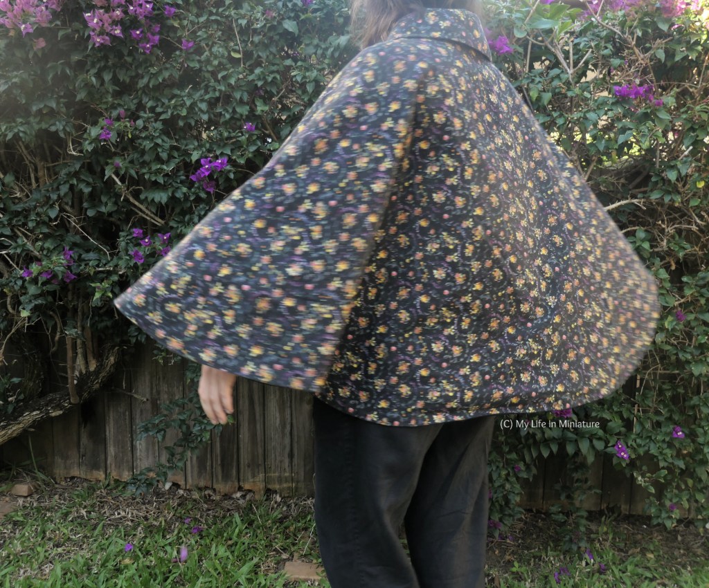 The author stands against a hedge, photographed from neck to knees. She is spinning in place, the cape flaring out slightly as she does so.