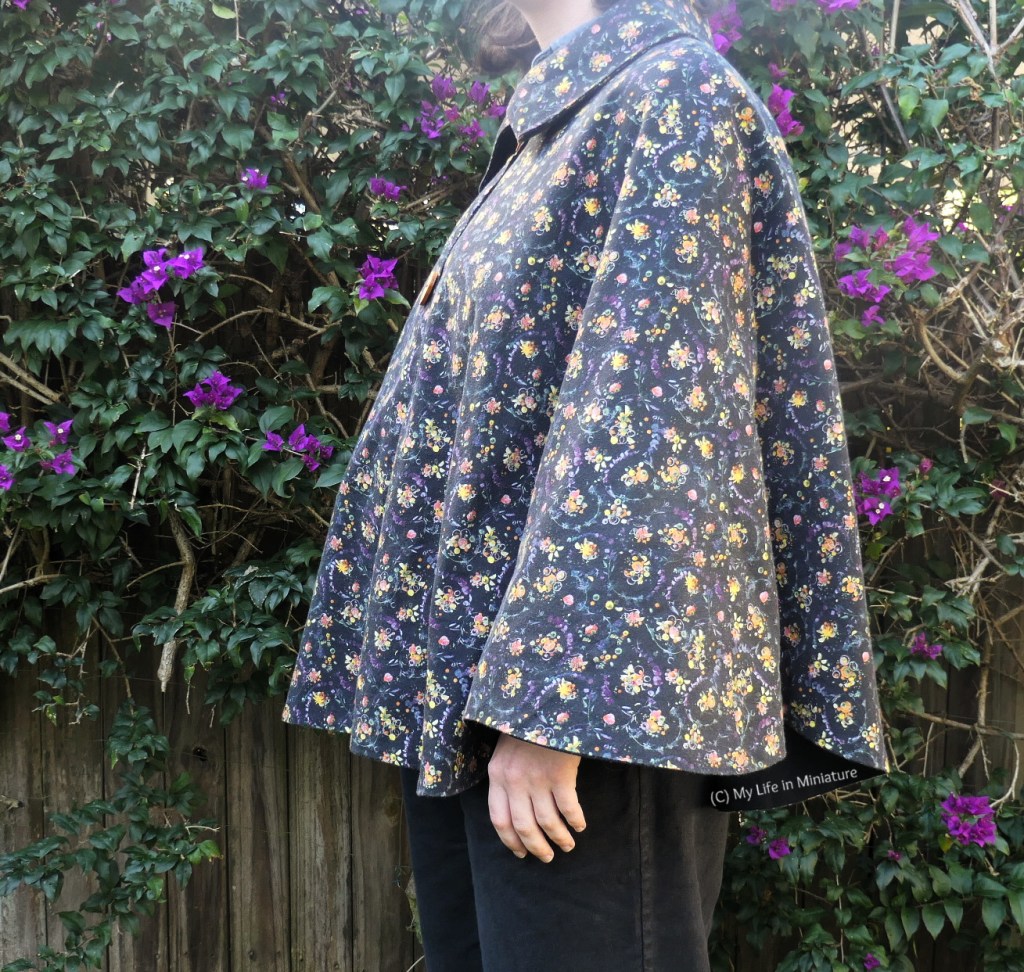 The author stands against a hedge, photographed from neck to thighs. She is side-on to the camera, with her arms hanging at her sides. The cape hem cuts off at her wrist, so her hand can be seen against her black pants.