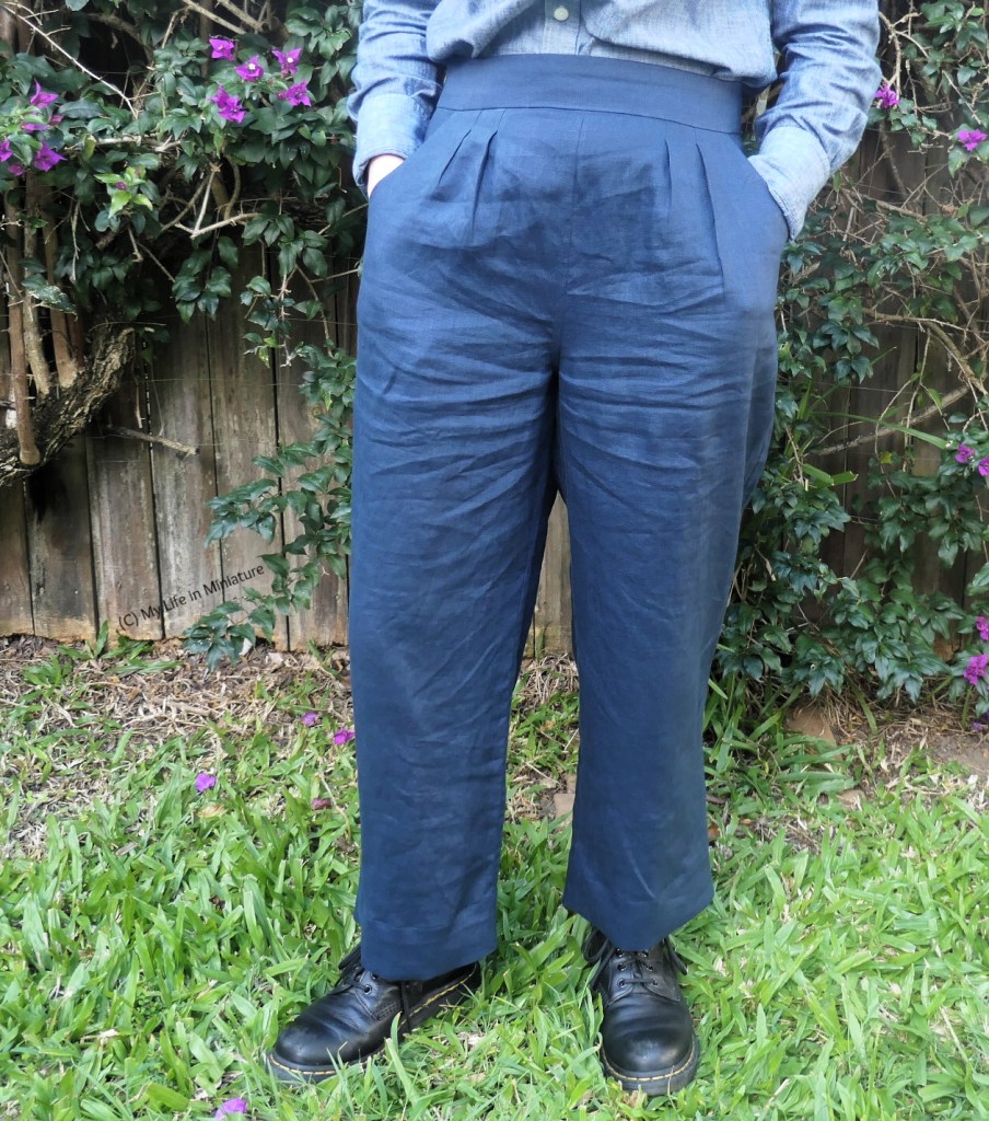 The author stands outside, against a hedge with small purple flowers. She's photographed from the waist down. She wears a pair of navy linen pants with a chambray blue shirt and black boots. She has ehr hands in the pants' pockets. 