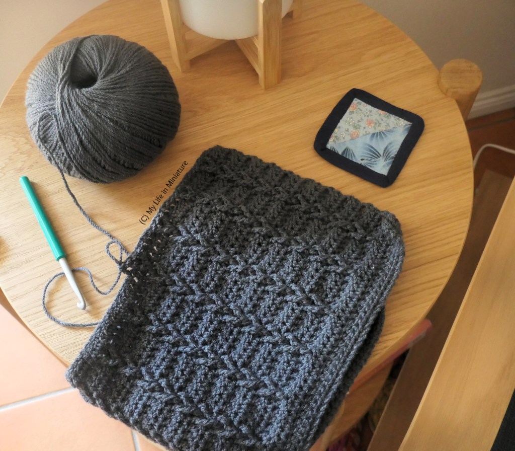 The coaster is on the small side table, with the lamp and a crochet project. The project is mid-row; the hook is resting nearby along with the ball of yarn.