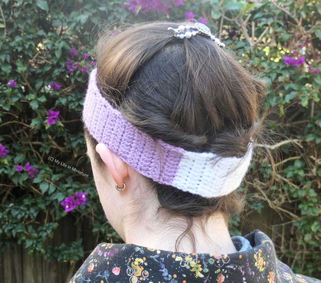 Back view of the author's head, taken outside. She wears a purple crochet headband around her head, and has her hair tucked into it at the back. This is so the white section of the purple headband at the base of her neck is visible. 