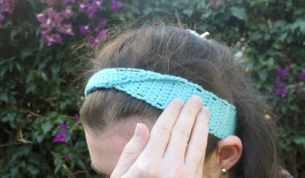 Close-up shot of the author's head, taken outside. She wears a light blue crocheted headband around her head, and has one hand up adjusting it. The headband has a twist in the centre front. 
