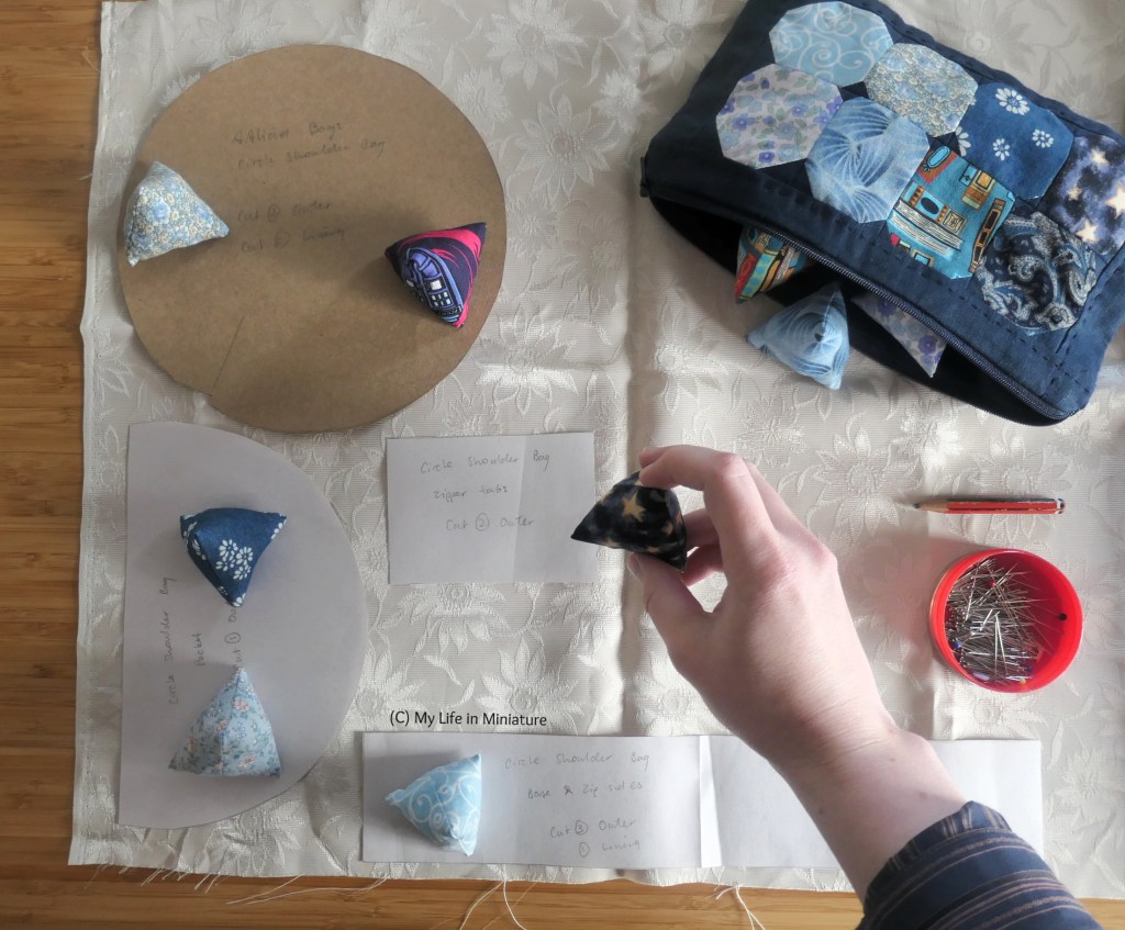 Overhead shot of the author's hand above fabric laid out on a wood surface. On the fabric (which is beige) are pattern pieces for a small bag, weighed down by the pattern weights. The author's hand reaches out with a weight to weigh down a pattern piece. To the right of her hand is the pouch, a pencil, and a pin tin.