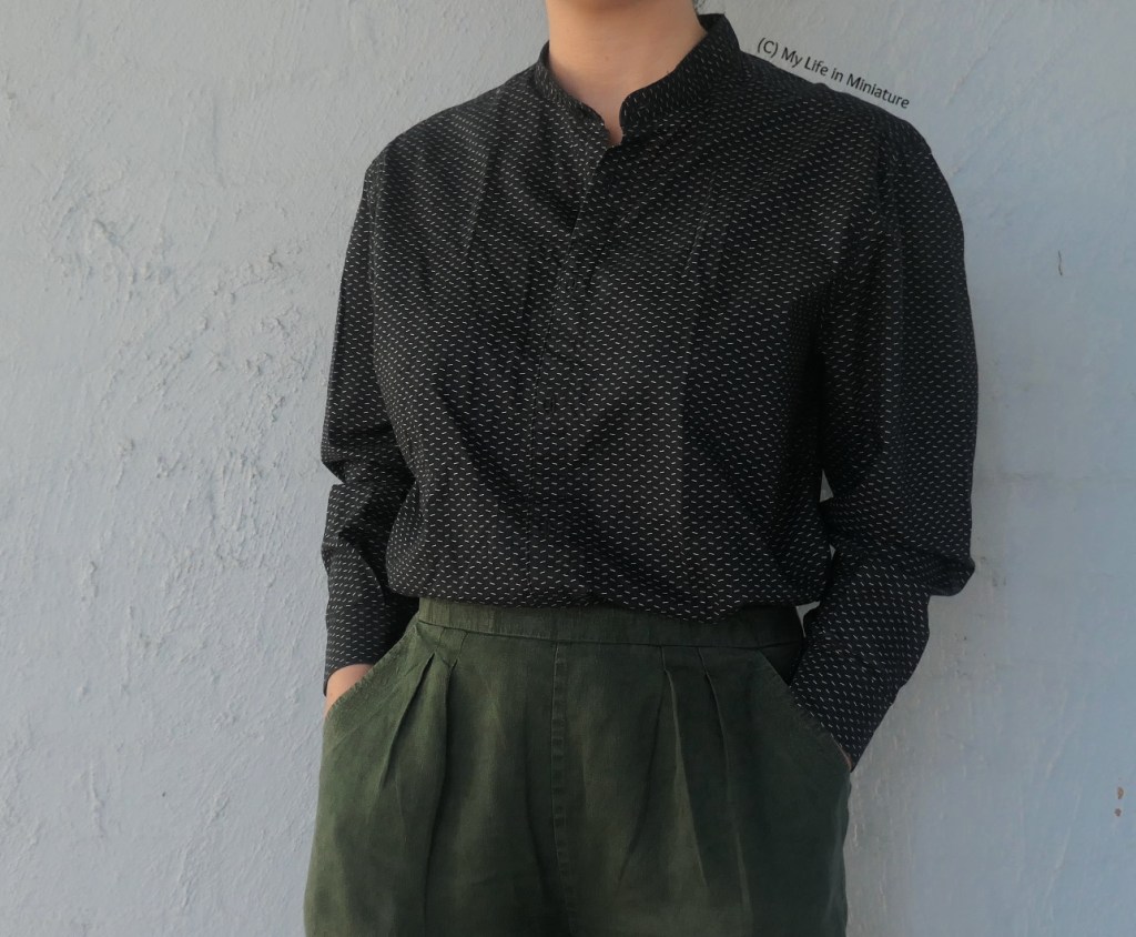 The author wears the black shirt with the white specks with dark green pants against a pale blue background. She's photographed from neck to thighs, and has her hands in her pockets.