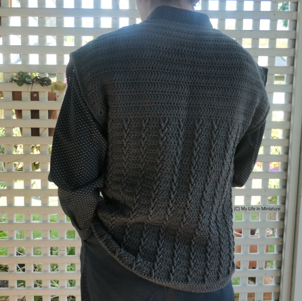 The author stands in front of a white lattice, photogrpahed from neck to thighs. She wears the crocheted tunic/button down shirt/navy pants combo, and is facing away from the camera so the back of the tunic is visible. 