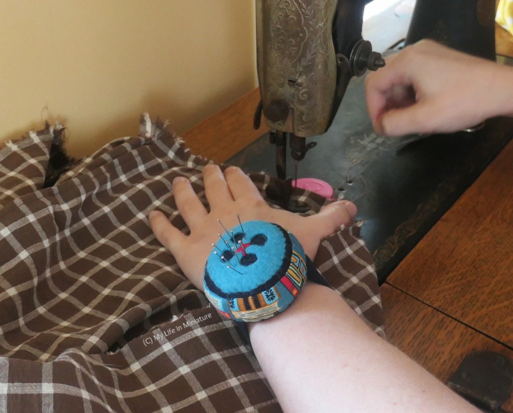 The author is sewing at an old sewing machine, photographed with only her lower arms visible. She wears the pincushion on the hand closest to the camera, and is in the middle of sewing a seam on a garment in progress. The pincushion has some pins in it. 