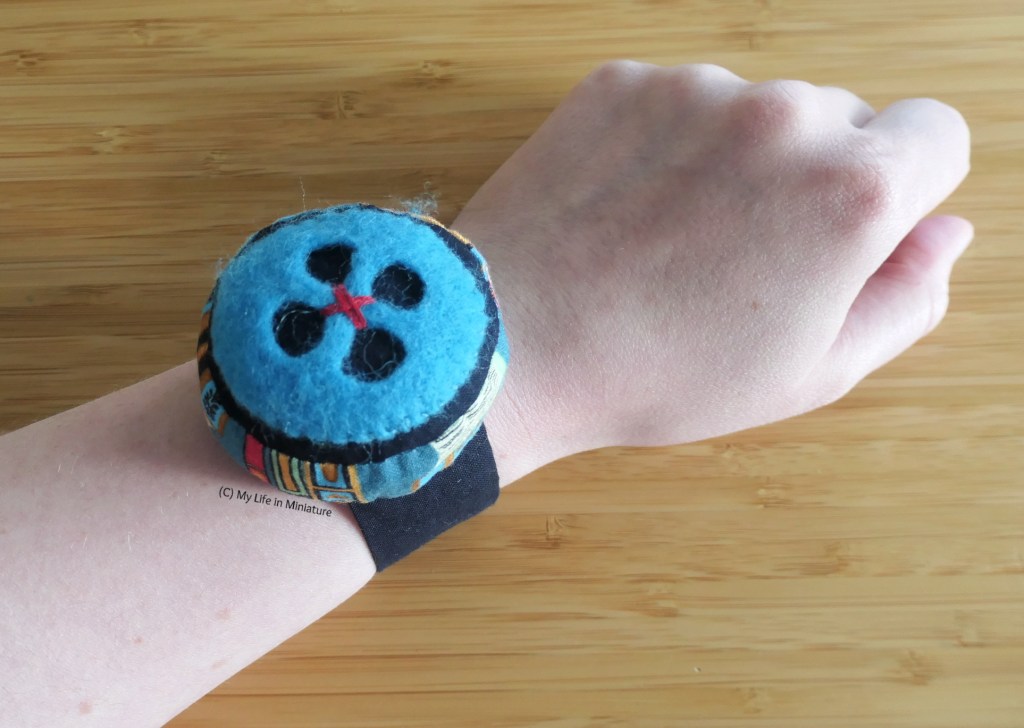 The author's wrist is photographed against a wood background, wearing the wrist pincushion. The button is clearer now, and the red stitches making a cross in the middle are visible. The sides of the pincushion are also more visible; they are made of blue fabric with books printed on. 