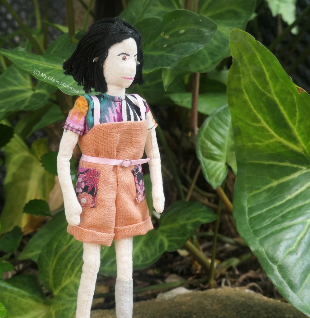 Tiffany stands in front of a background of leaves and stems, looking off to the right of the frame. She has her hands by her sides and smiles softly. She still wears the dungarees and shirt combination.