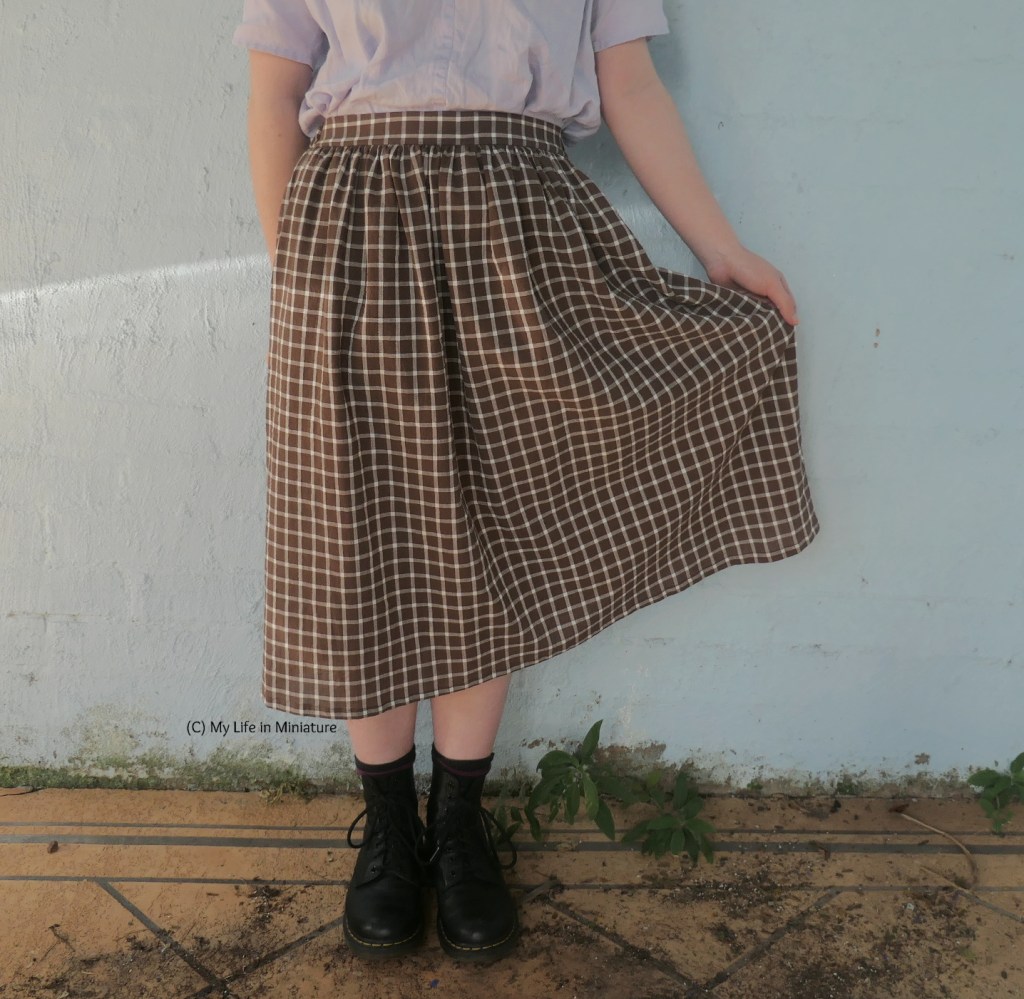 The author stands in front of a blue wall, photographed from waist to feet. She wears the same brown, gathered skirt with purple top and boots. She holds the skirt out with one hand to show the print, which is white gingham stripes on a brown background.