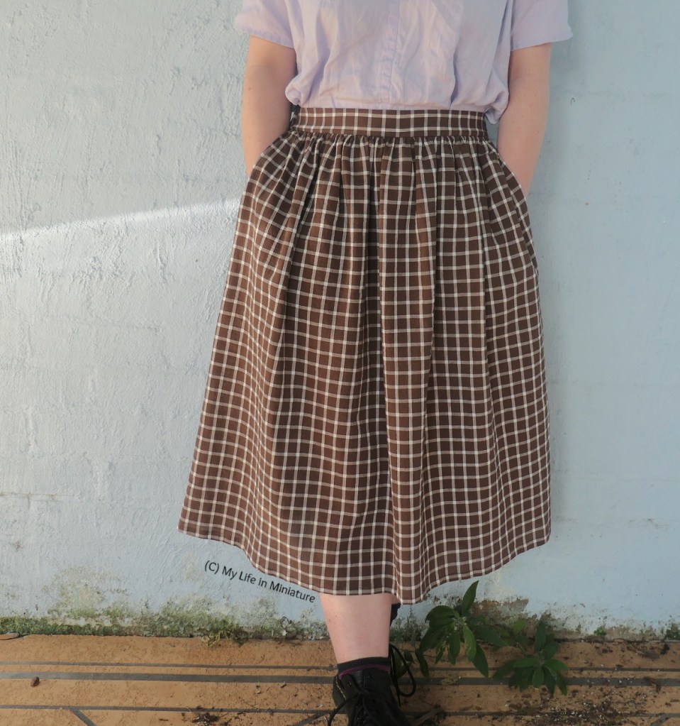 The author stands in front of a blue wall, wearing the brown skirt. Photographed from waist to feet, she has both hands in the skirt's side seam pockets. 