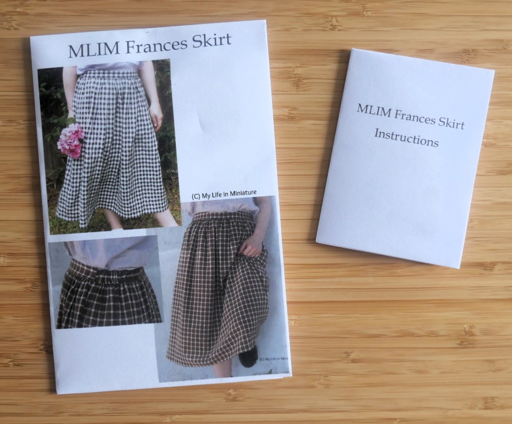 A sewing pattern pouch for the 'MLIM Frances Skirt' and a corresponding instruction booklet are laid flat on a wood surface. 