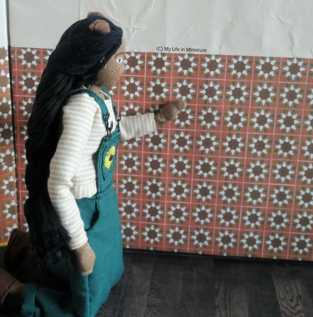 Petra kneels near the base of one wall in Tea at Yaz's, running a hand over the tiles. They have an eight-pointed star design, dark brown centre with white arms to the stars. There are dark green leaves inbetween the white arms of the stars, all on a terracotta background.