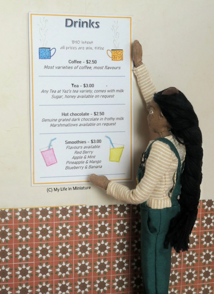 Close-up of Petra hanging the drinks menu back up on the wall of Tea at Yaz's, where it went beforehand. The drinks menu looks the same as before, with coffee, tea, hot chocolate, and smoothies available. 