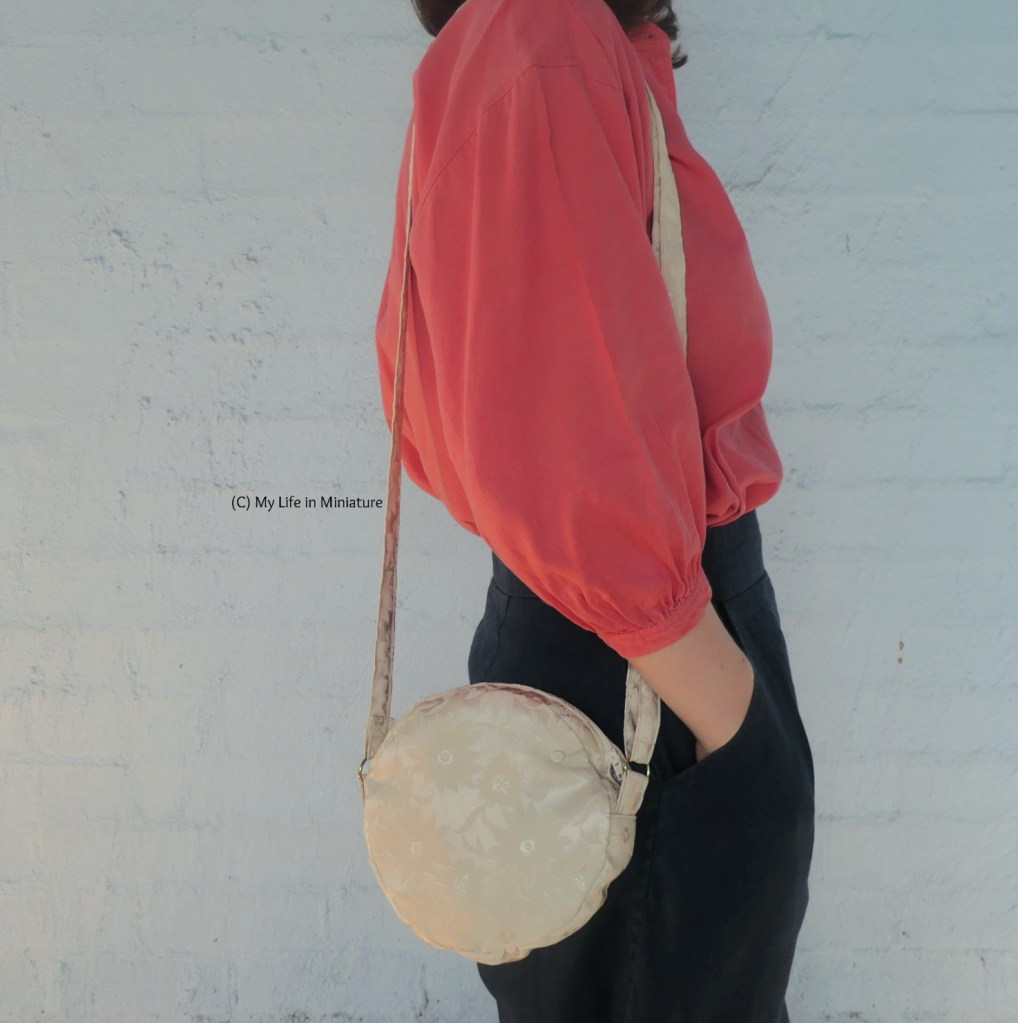 The author stands in front of a pale blue wall, wearing a peach top with navy pants. She is side-on to the camera, with hands in her pockets. Over her shoulder is a small, cream-coloured, circular bag.