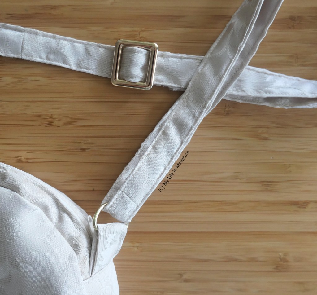 Close-up of the small cream bag. One of the sides of the bag is visible, showing the gold D-ring and the strap that loops into it. Also visible is a gold sliding buckle further up on the strap. 