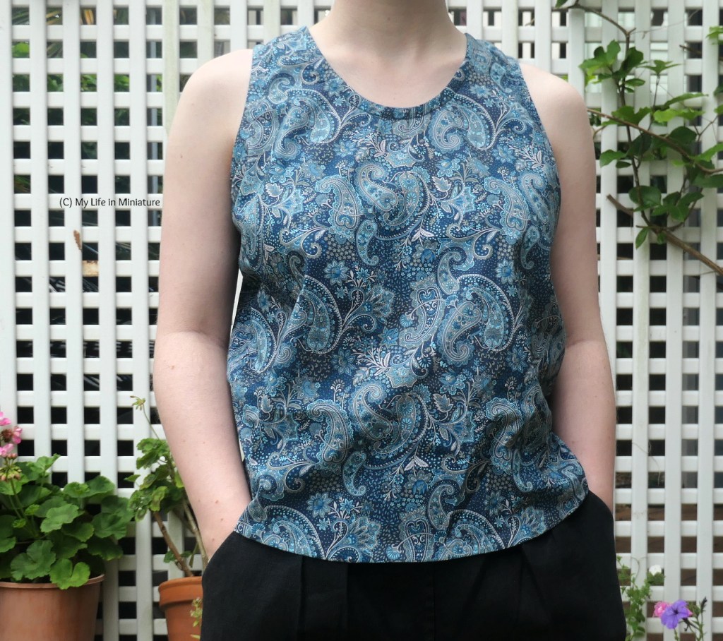 The author stands in front of a white lattice, which has a vine growing up it. She wears the same blue paisley singlet top and black pants as before. She has her hands in her pockets, and is photographed from neck to hips.