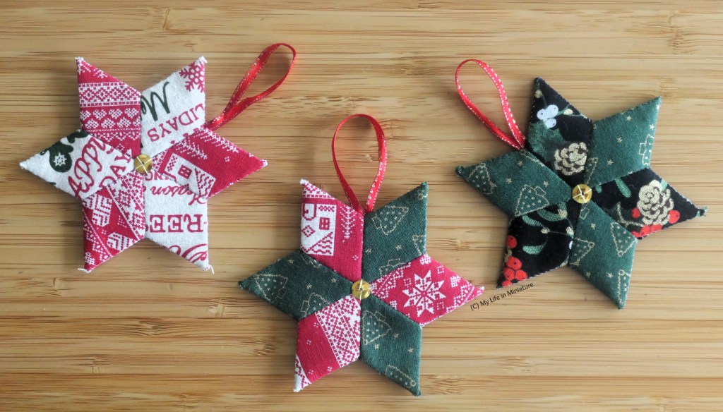 Three star-shaped Christmas decorations are laid out on a wood surface. They are each made of six diamonds of Christmas fabric, sewn into star shapes. They all have red ribbon as hanging loops, and a gold sequin sewn in the centre. From left to right, the colour schemes are: white and red, red and green, and green and black. 