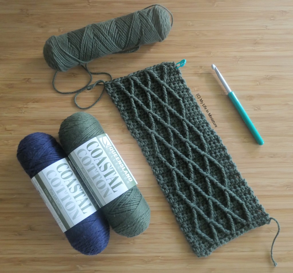 Overhead shot of a piece of crochet laid flat on a wood surface. The crochet is in a dark green yarn, and has raised criss-cross patterns on it. Next to the crochet is the crochet hook used, the yarn ball being used, and two of the other yarn balls being used for the project. 
