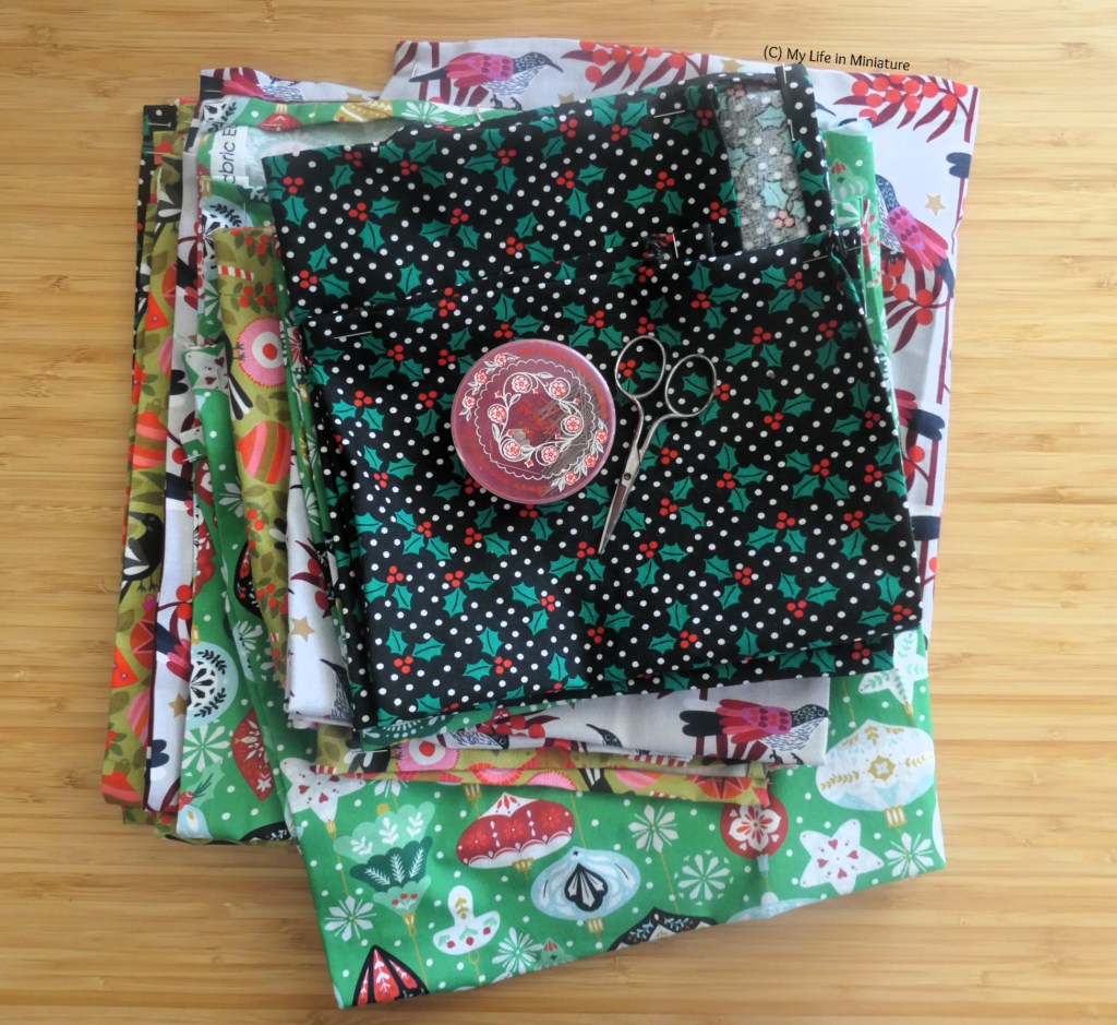 A stack of Christmassy fabric is photographed from above on a wood surface. At the very top of the pile is a red pin tin and a pair of embroidery scissors. 