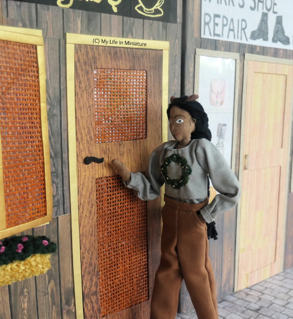 Petra walks towards the front door to Tea at Yaz's, reaching out for the doorhandle. She wears a grey long-sleeved shirt with a wreath embroidered on the front, with brown pants. 