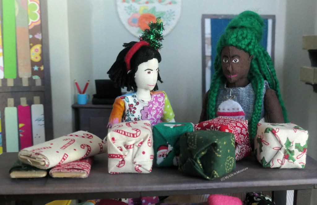 Tiffany and Hazel stand at the cutting table in Needle & Thread. On the table are five decorative presents, freshly wrapped in Christmassy fabric. The leftover bolts are set to the side. 