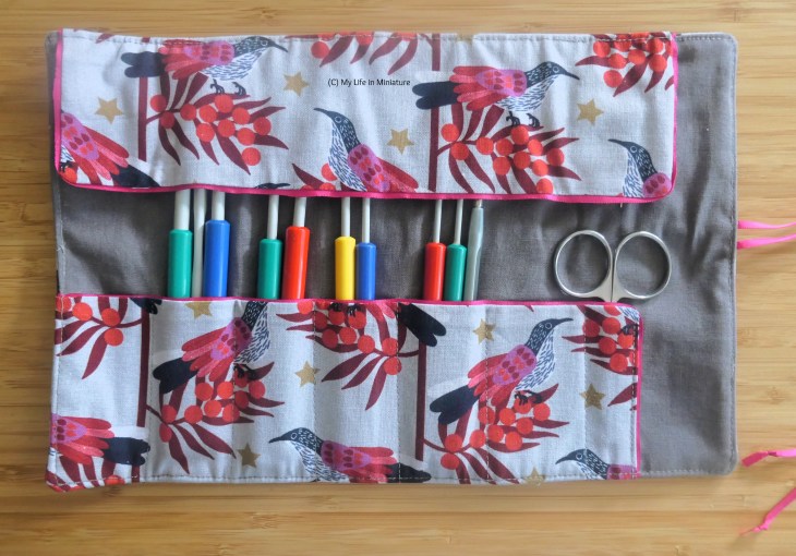 A hook roll is unrolled on a flat surface, photographed from above. It contains a lot of hooks, and a pair of scissors. The lining fabric is grey, and the outer fabric is light grey with birds, red foliage, and small gold stars on. 