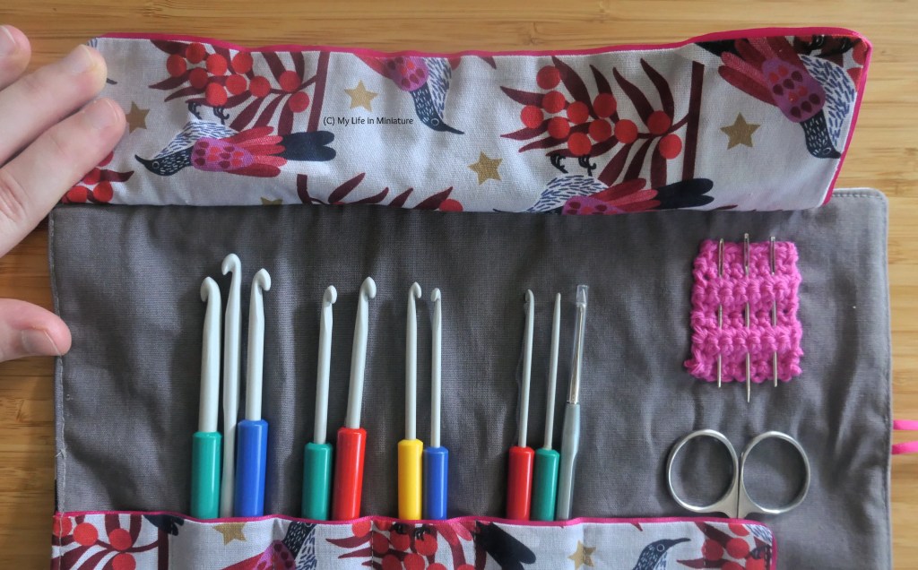Close-up of the underside of the flap at the top of the hook roll, which is being held up by the author's hand. The top half of the hooks are visible, as is a small square of hot pink crochet with three yarn needles poked through. The underside of the flap is the same bird fabric as the exterior.
