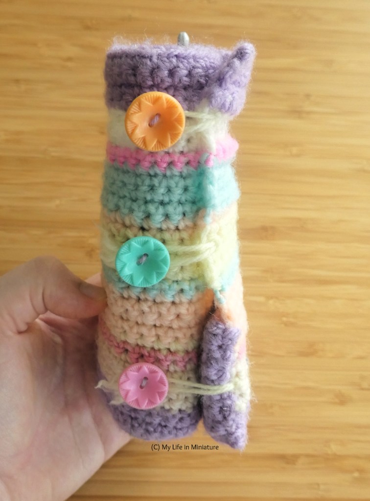 The author's hand holds her old hook roll over a wood background. The hook roll is rolled up, secured with three buttons. It is crocheted in a variety of pastel shades. 